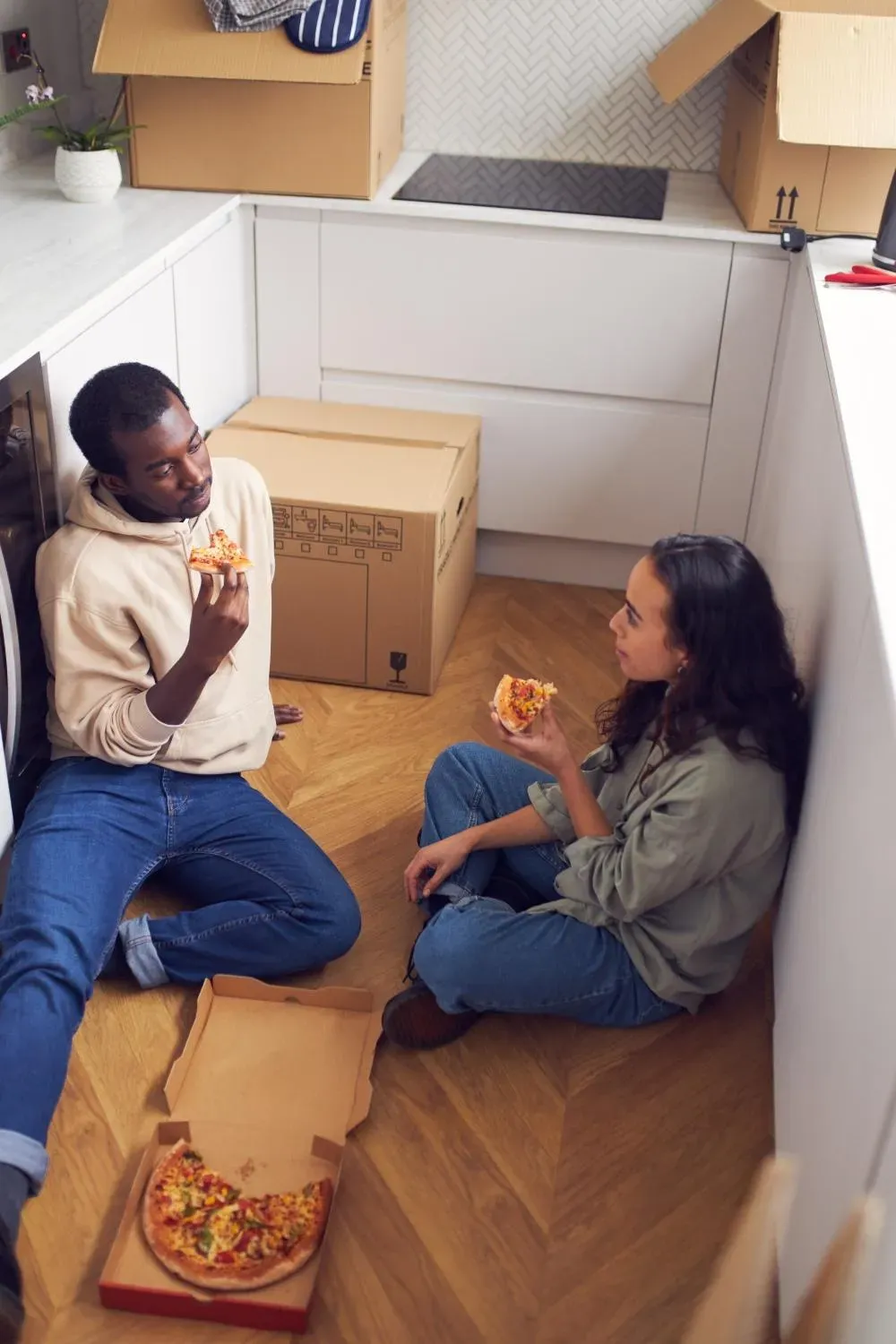 Två personer sitter på golvet och äter pizza i tomt rum med flyttkartonger.
