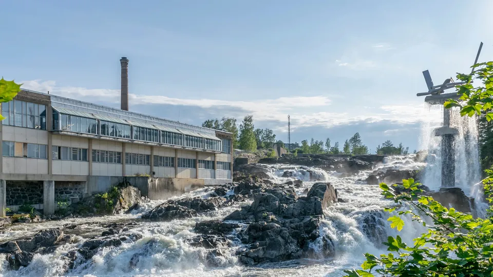 Foss som fosser over steiner ved siden av industribygg med murstein skorstein.