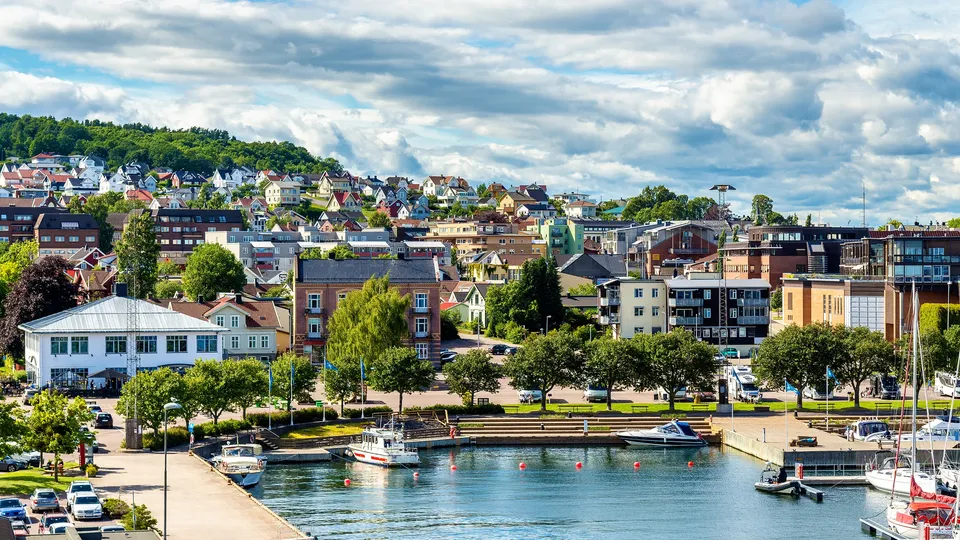Kystby med fargerike bygninger, marina med seilbåter og skogkledd bakke.