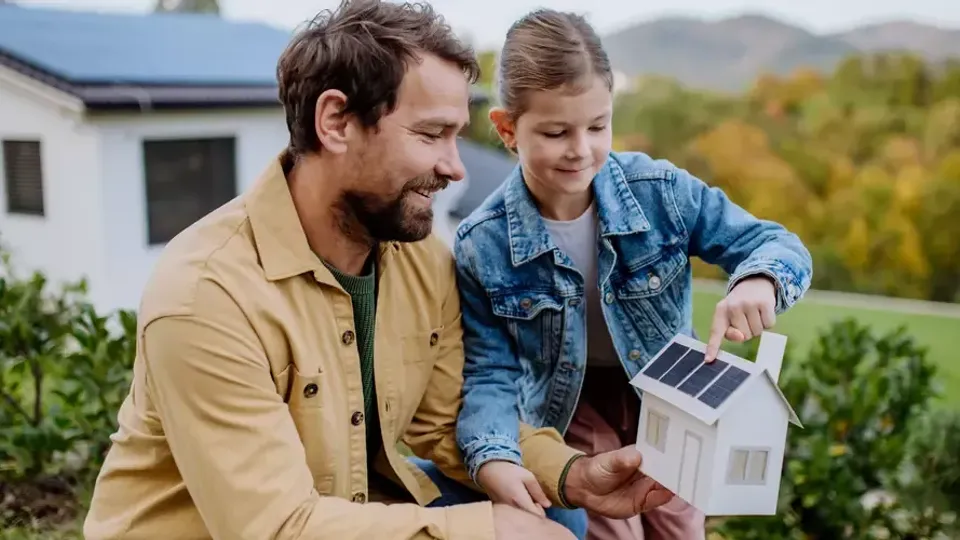 En man och ett barn sitter utomhus och tittar på en modell av ett hus med solpanel.