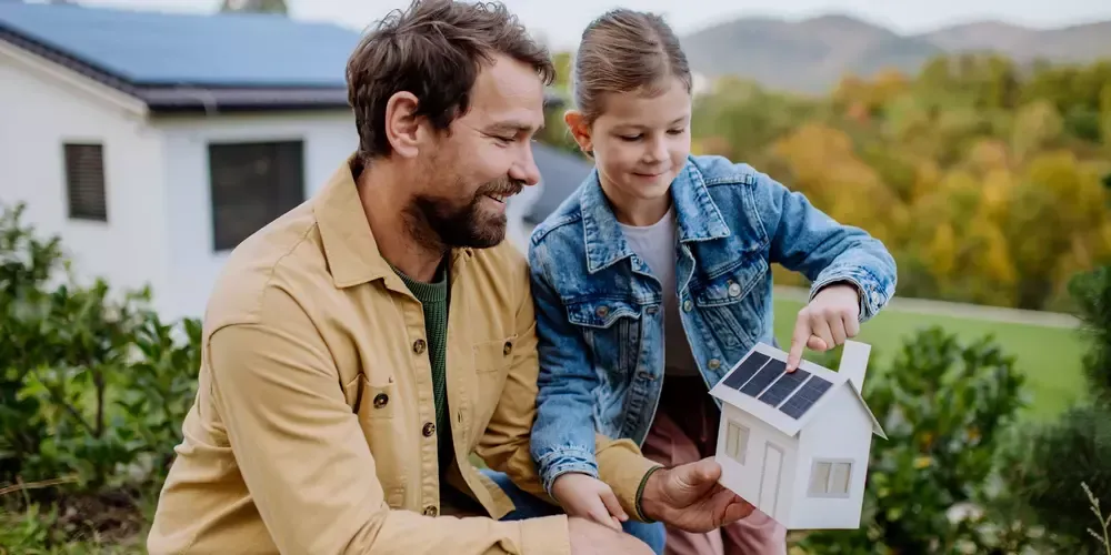 En man och ett barn sitter utomhus och tittar på en modell av ett hus med solpanel.