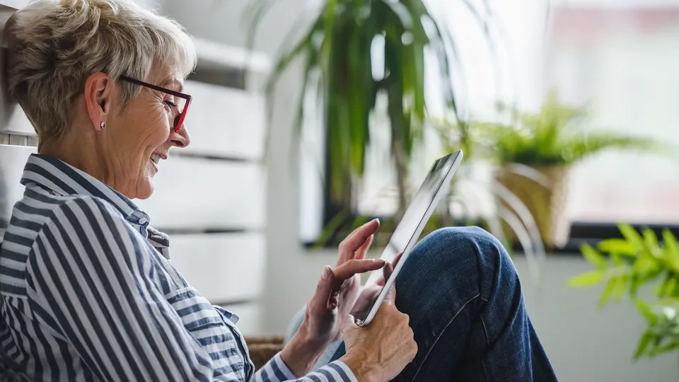 Ældre kvinde med briller bruger tablet, mens hun sidder indendørs.