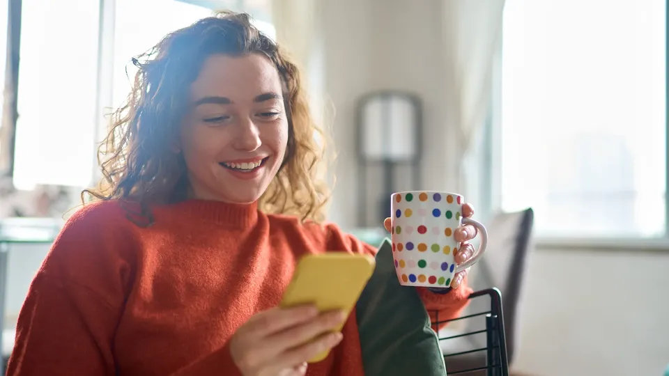 Kvinde smiler mens hun holder farverig prikket kop og ser på gul telefon.