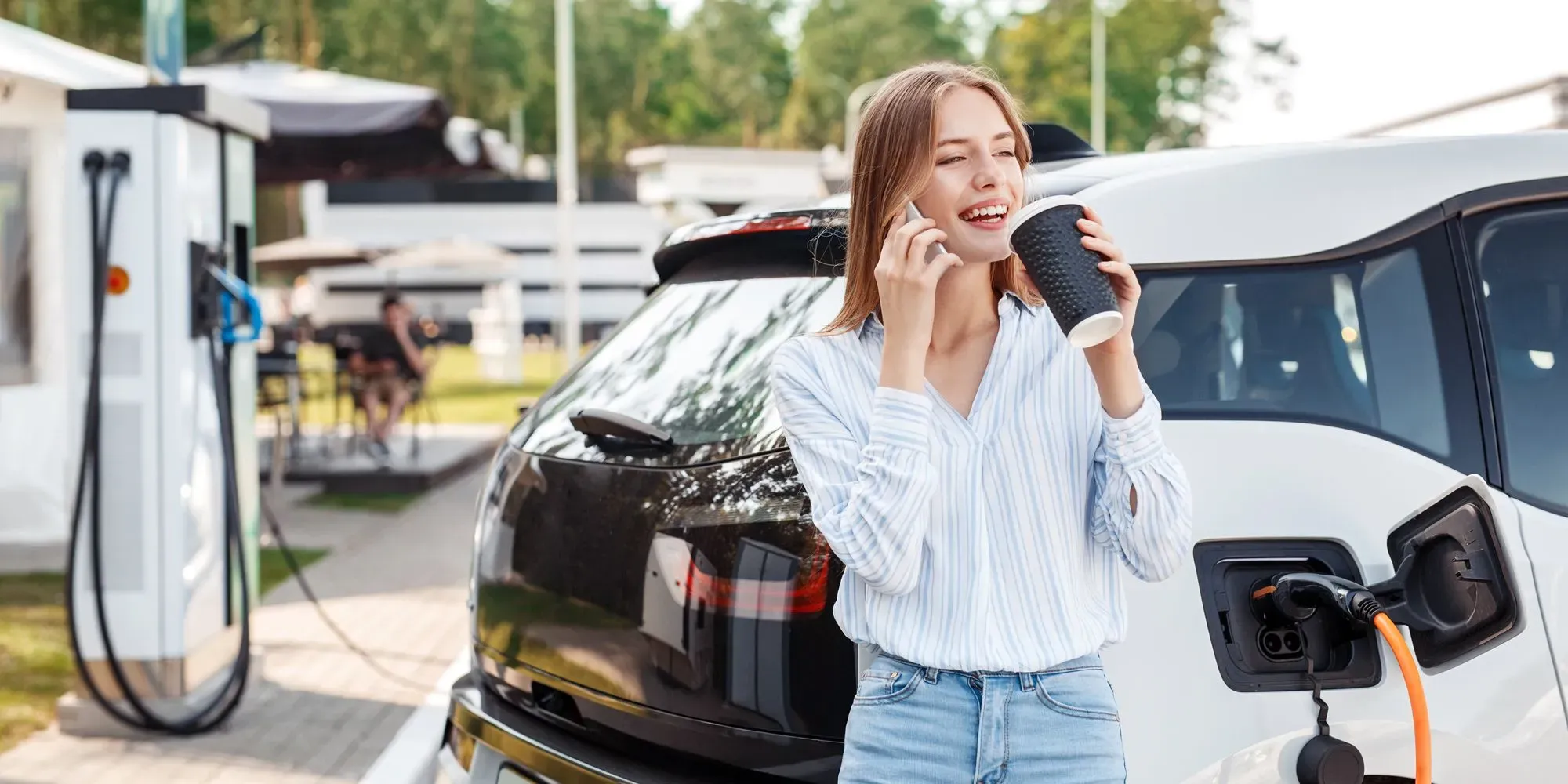 Bilde av en dame med kaffekopp som tar pause mens bilen lades i bakgrunnen