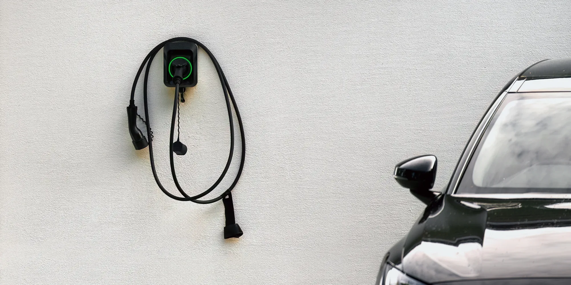 Elektrisk køretøjsopladningsstation monteret på hvid væg med sort kabel rullet op nedenfor.