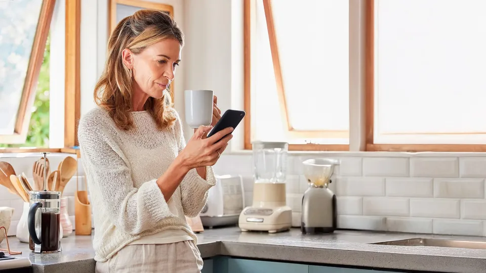 Kvinde i køkken holder kaffekop og tjekker smartphone.