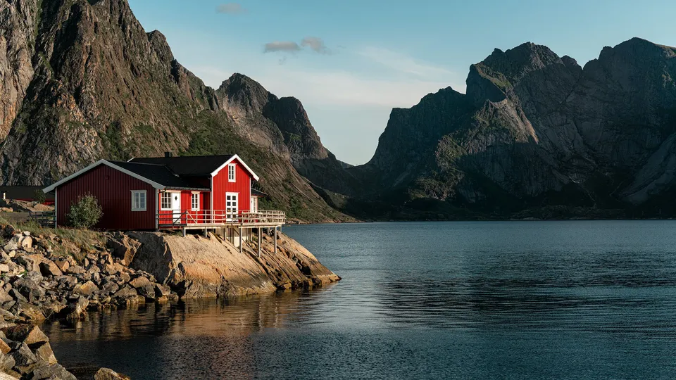 Rød hytte på påler ved en fjord omgitt av bratte fjell.