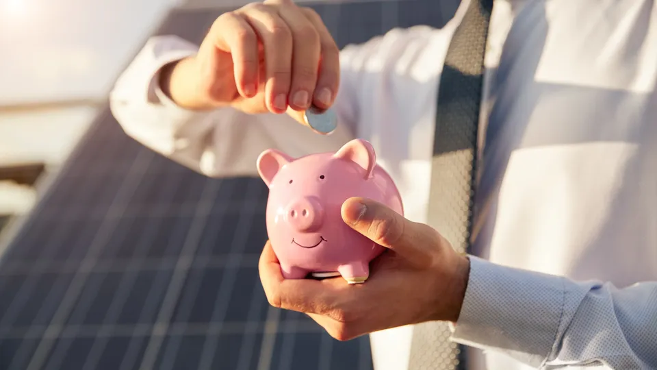 Person steckt eine Münze in ein Sparschwein vor einer Photovoltaikanlage – Symbolbild für Photovoltaik-Kosten und Einsparungen