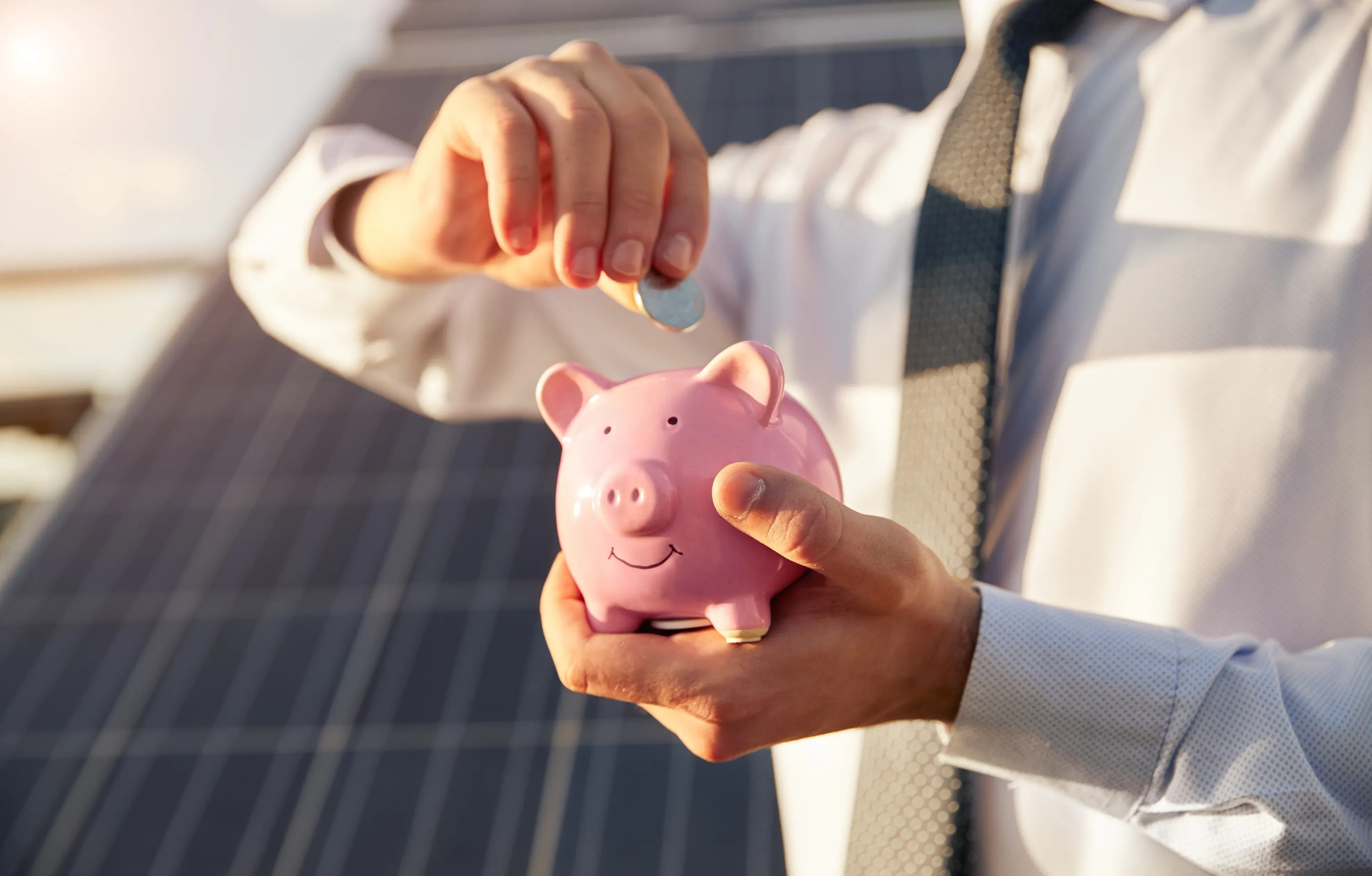 Person steckt eine Münze in ein Sparschwein vor einer Photovoltaikanlage – Symbolbild für Photovoltaik-Kosten und Einsparungen