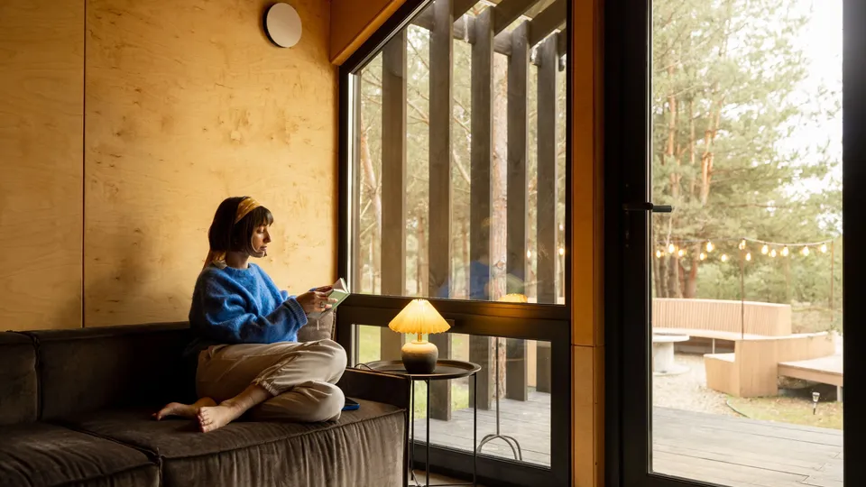 Kvinne sitter på sofa ved siden av et stort vindu i sitt moderne arkitekt-designede hus.