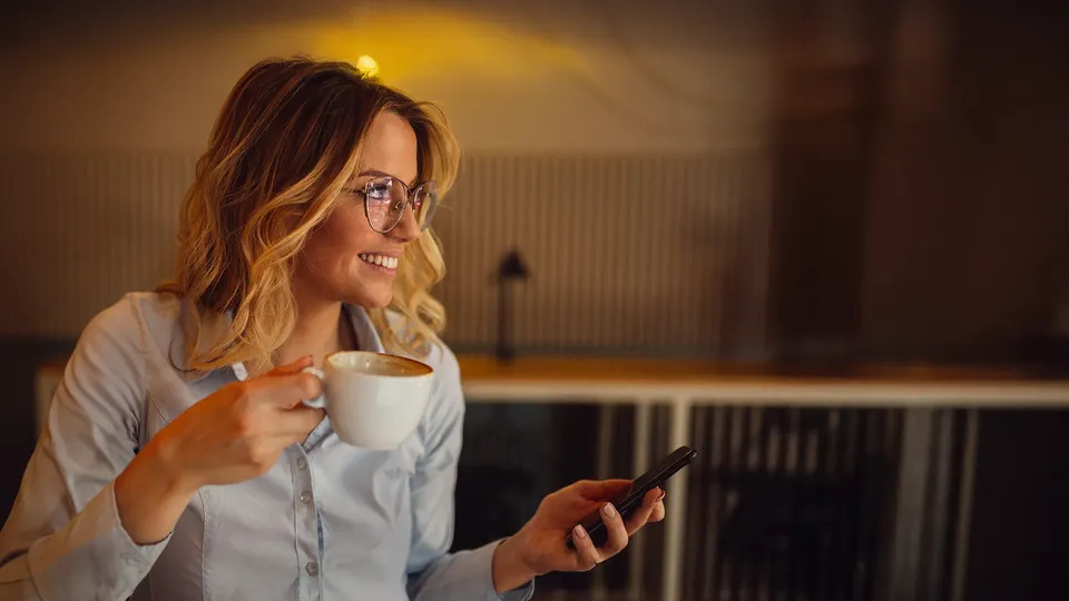 Kvinde holder kaffekop og smartphone, smiler mens hun ser væk.