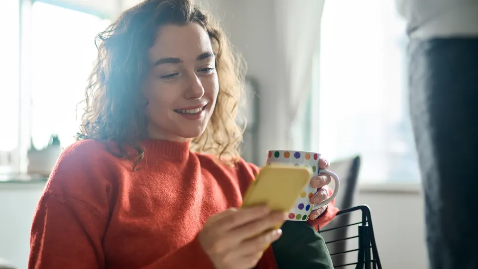 Kvinde smiler mens hun kigger på telefon og holder farverig kop.
