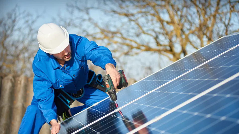 Trabajador en uniforme azul instalando panel solar con taladro.