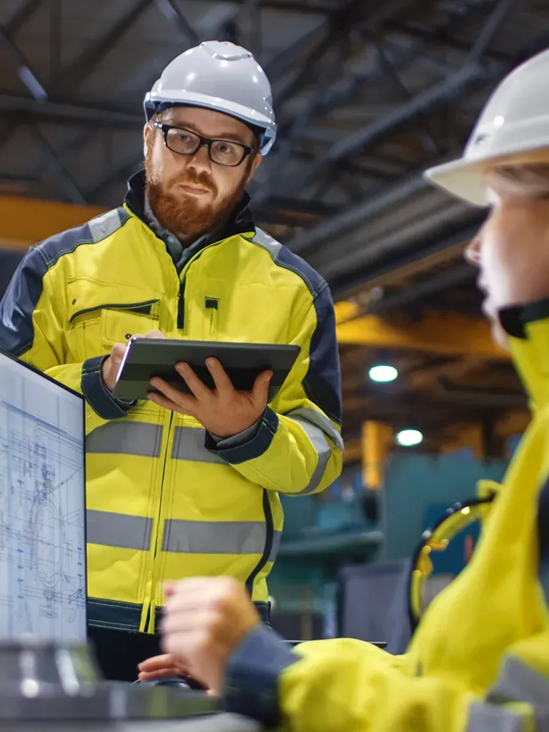 Trabajador de construcción sostiene tableta y discute planos con colega.