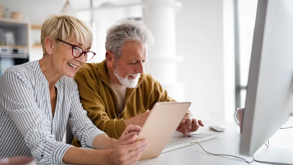 To ældre voksne ser på en tablet sammen ved et skrivebord.