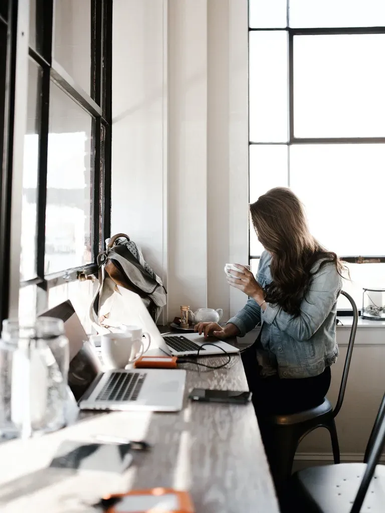 Mujer trabajando en escritorio con portátil junto a ventana brillante.