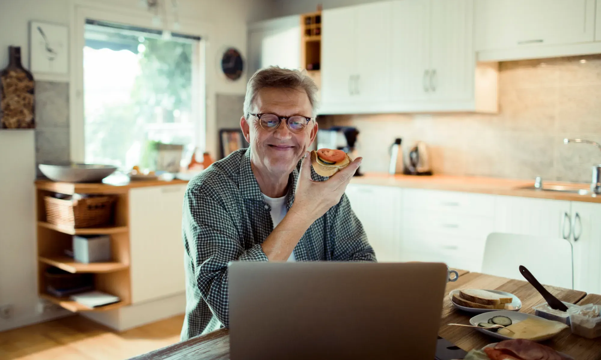 En man äter en frukostmacka vid köksbordet och ler mot en öppen laptop.
