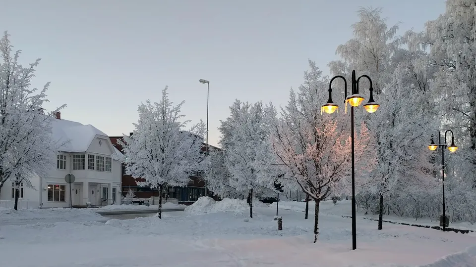 Snøfylt torv med rimfrostkledde trær og tente lyktestolper i skumringen.