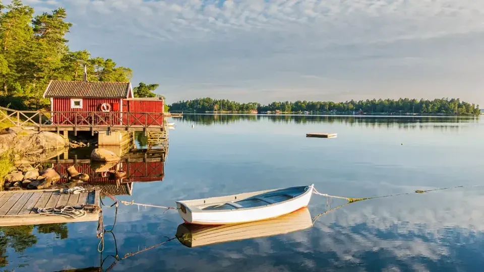 Liten röd stuga vid sjökanten med båt och spegelblankt vatten en klar sommardag i Sverige.