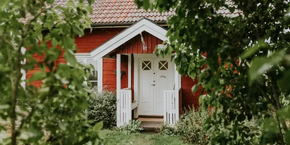 Ett rött hus med vita knutar och lummigt av björk.