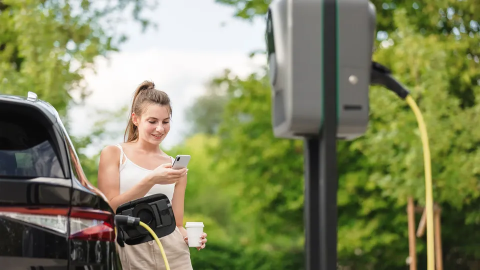 Kvinde bruger smartphone mens hun oplader elektrisk køretøj ved en ladestation.