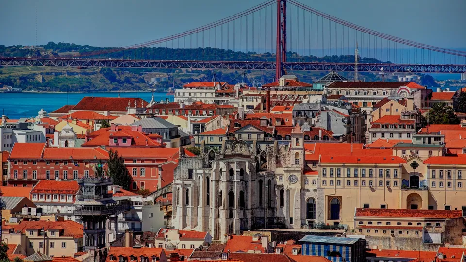Paisagem urbana de Lisboa com edifícios de telhados vermelhos, igreja e ponte suspensa sobre água azul.