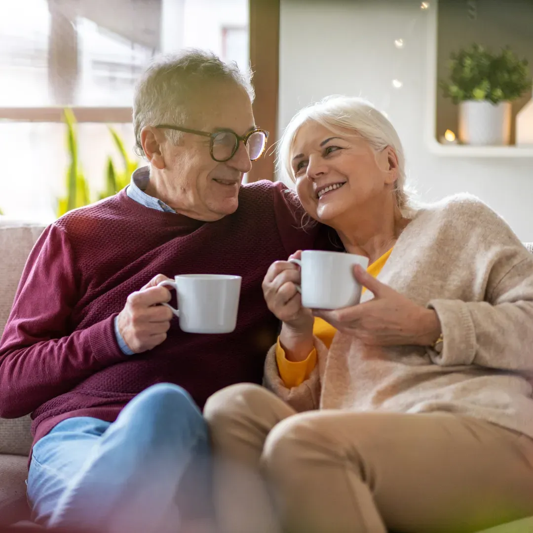 Hombre y mujer mayores sentados en sofá sosteniendo tazas y sonriéndose.
