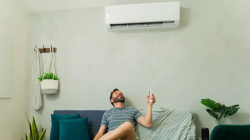 Mand på sofa holder fjernbetjening mod klimaanlæg på væg.