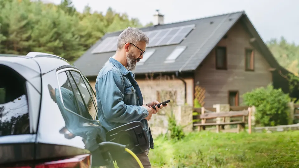 Mand holder telefon ved elbil ved hus med solpaneler.
