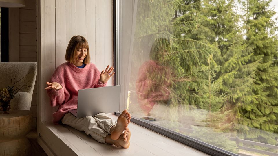 Bilde av en kvinne som sitter i en dyp vinduskarm på en laptop og smiler.