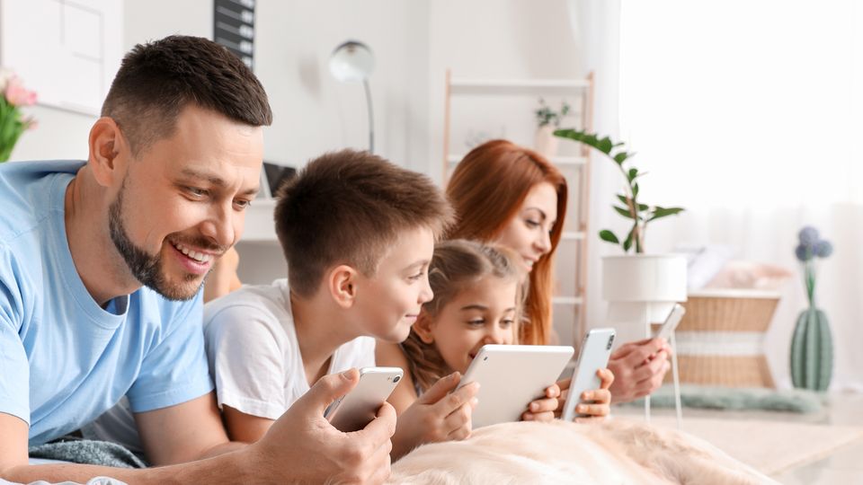 Bilde av en smilende familie som ligger på gulvet og ser på hver sin skjerm.
