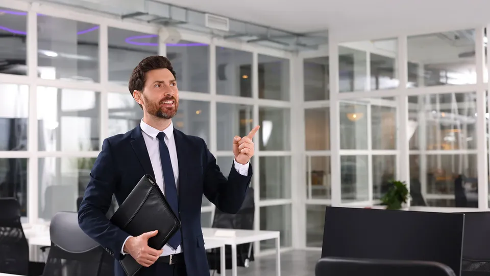 Smiling businessman in a dark suit and tie holding a leather portfolio, pointing upward while presenting a modern open-plan office space with large glass walls and bright lighting.