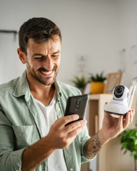 Man holding smartphone and security camera, smiling while viewing device.