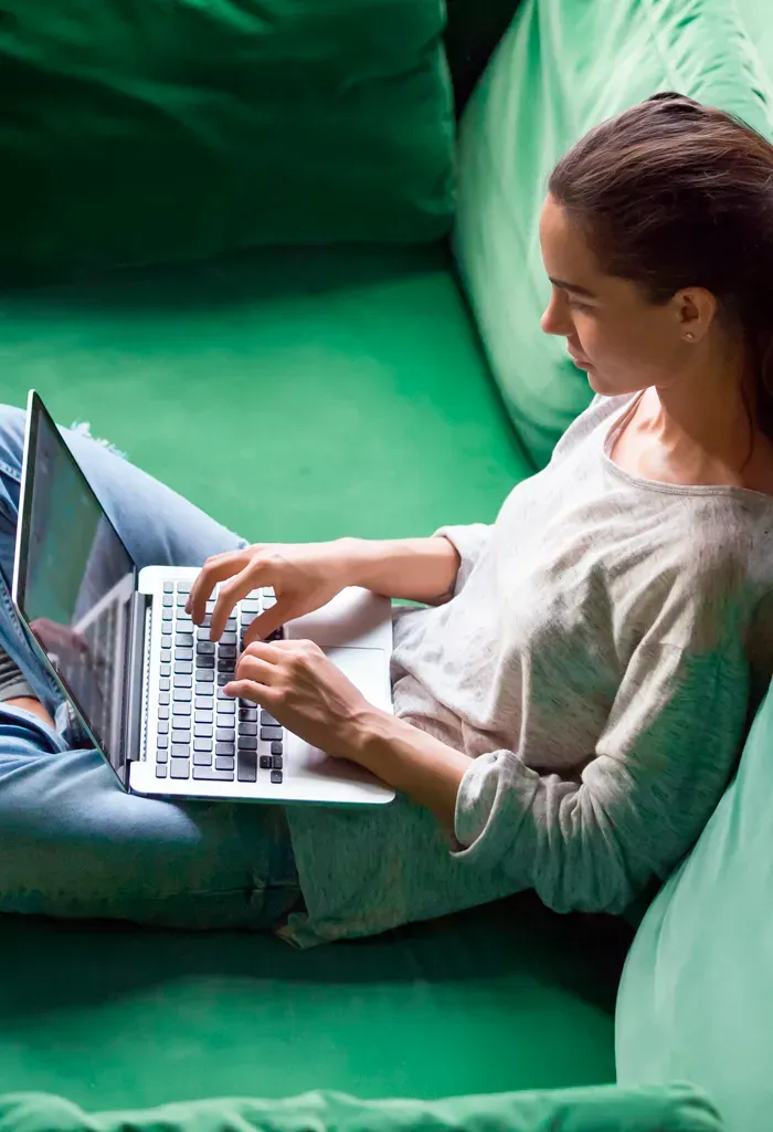 Mujer escribiendo en laptop mientras está sentada en cojines verdes.