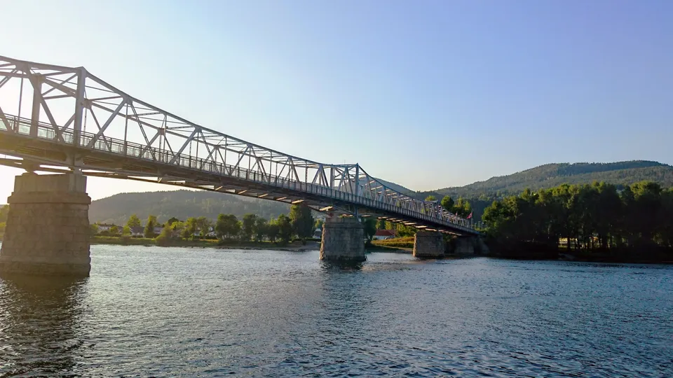 Stålgitterbridge som spenner over en elv med steinpilarers og skogkledde åser.
