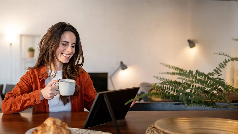 Kvinde holder kop og ser på tablet ved skrivebord.