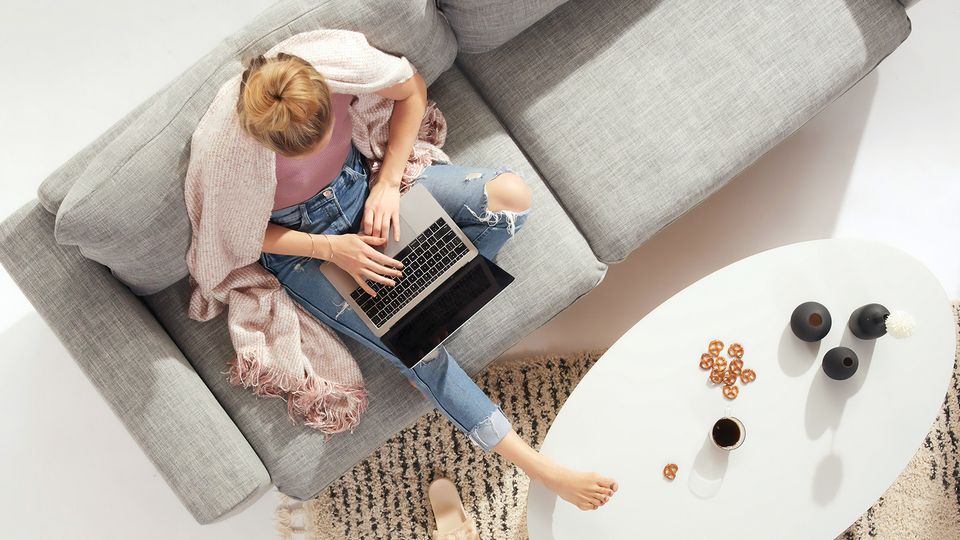 Bilde av en kvinne som skriver på en pc mens hun sitter i sofaen med et pledd over skuldrene.