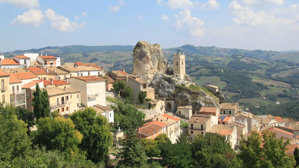 Villaggio medievale su collina con edifici in pietra e una grande formazione rocciosa con torre di chiesa.