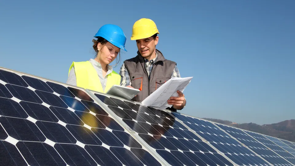 Two workers in hard hats reviewing blueprints on solar panels.