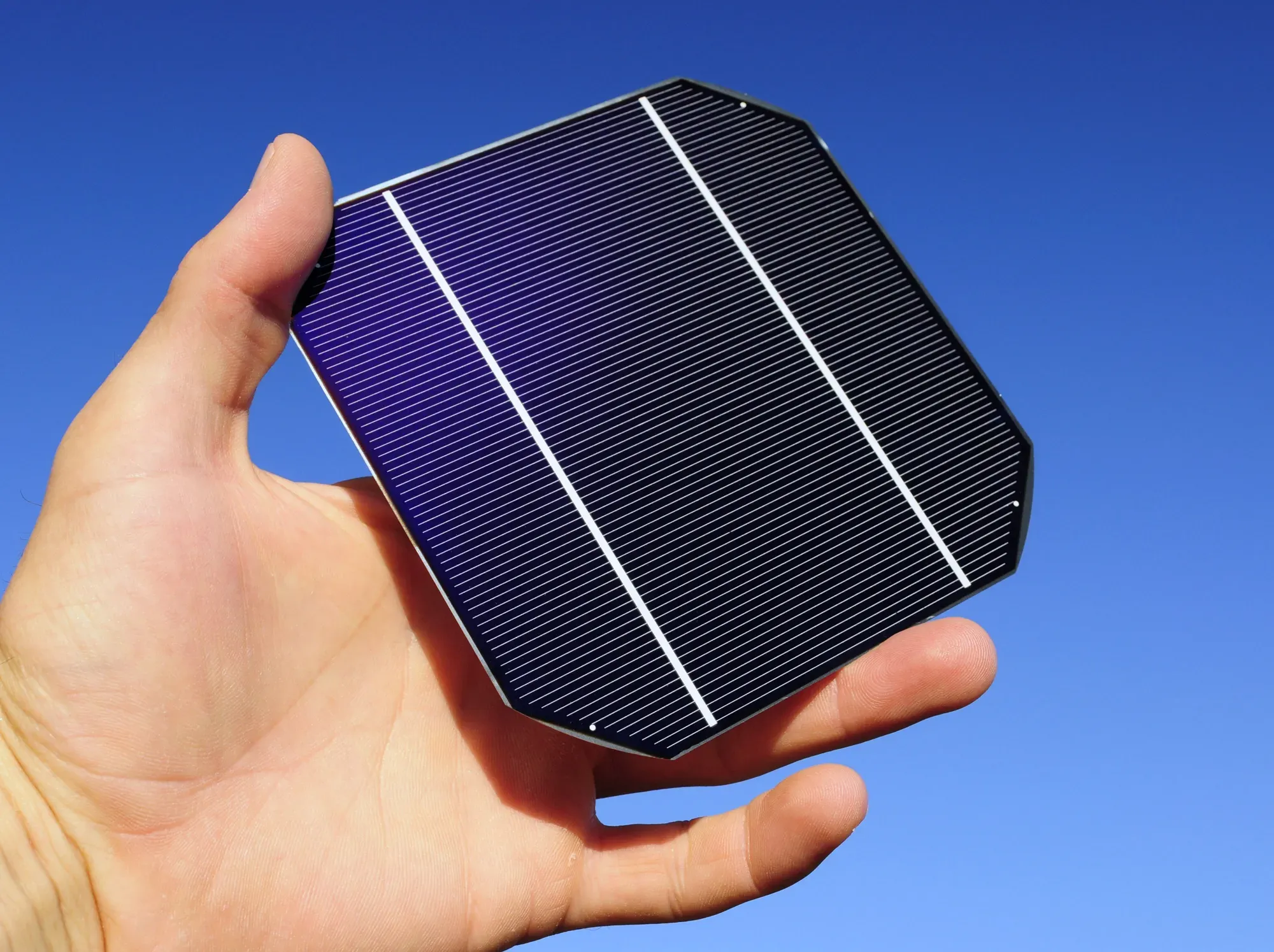 Hand hält eine Solarzelle mit blauer Oberfläche und weißen Gitterlinien vor blauem Himmel.