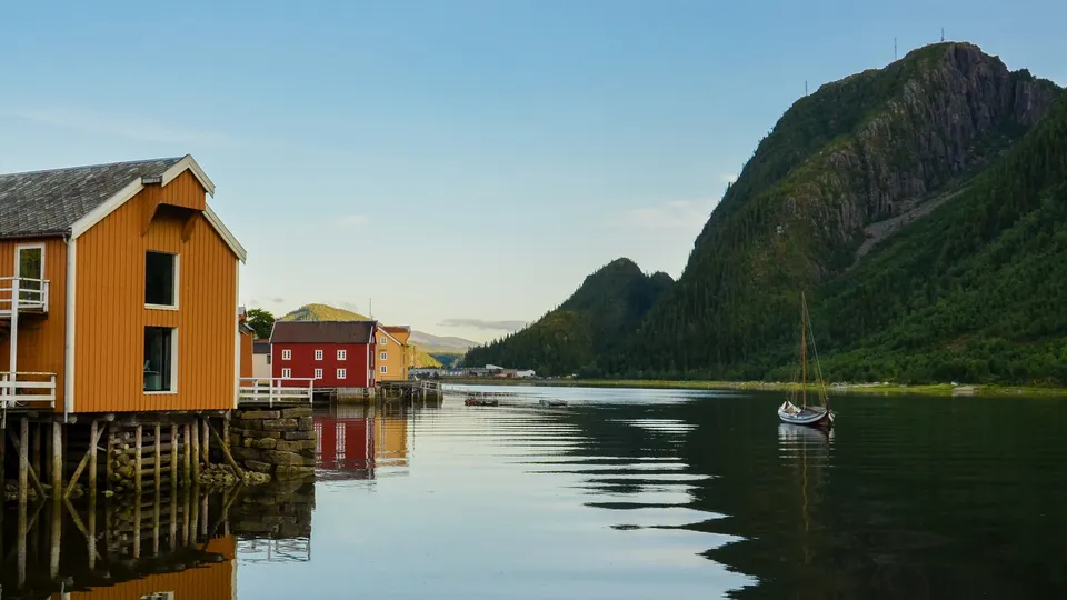 Fargefulle hus på påler ved en stille fjord med skogkledde fjell og en seilbåt.