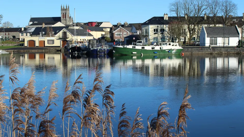 County Leitrim: Green and white boat moored on calm river with waterfront buildings and church spire.