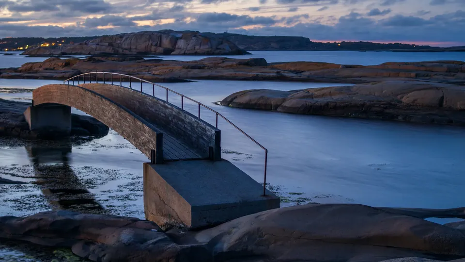 Buet trebrygge som strekker seg over steinete kystvannet ved skumring.
