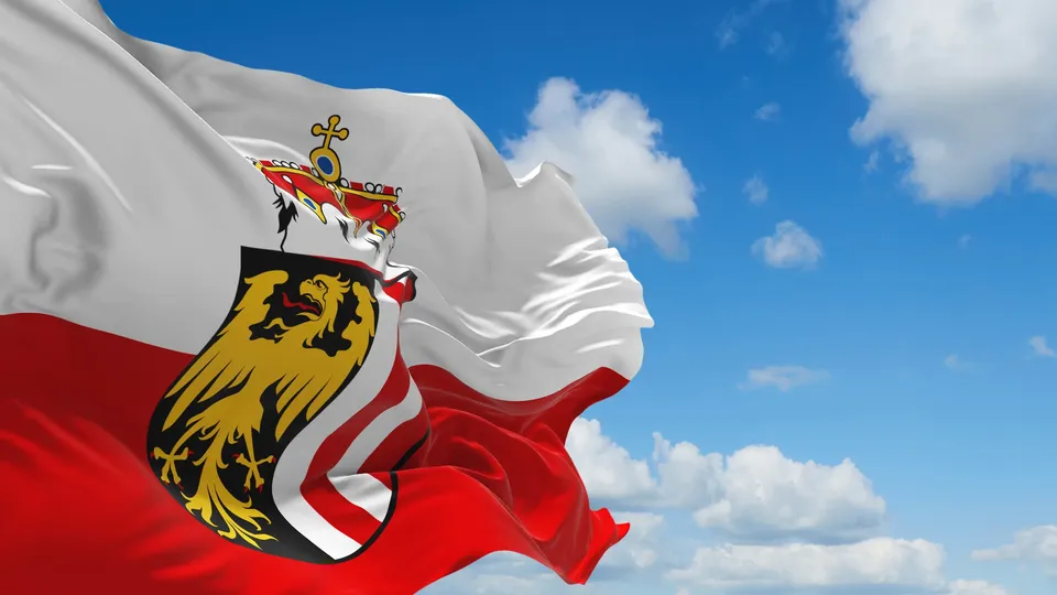 Rot-weiße Flagge Oberösterreichs mit dem oberösterreichischen Wappen vor blauem Himmel mit Wolken.