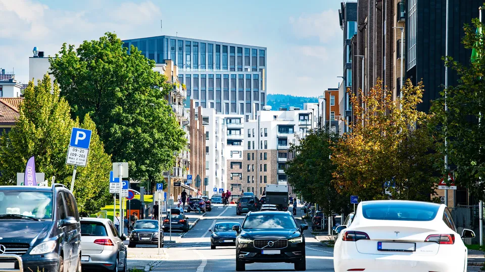 Urbant gatestrekk med parkerte og kjørende biler omgitt av trær og moderne bygninger.