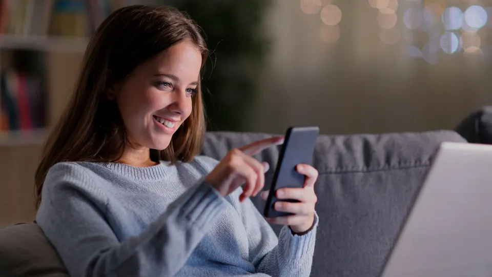 Mujer sonriendo mientras usa un smartphone en el sofá.