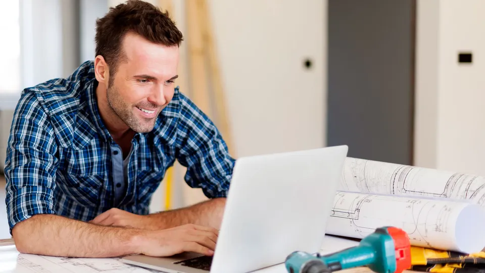 Hombre usando laptop en escritorio con planos de construcción y herramientas.