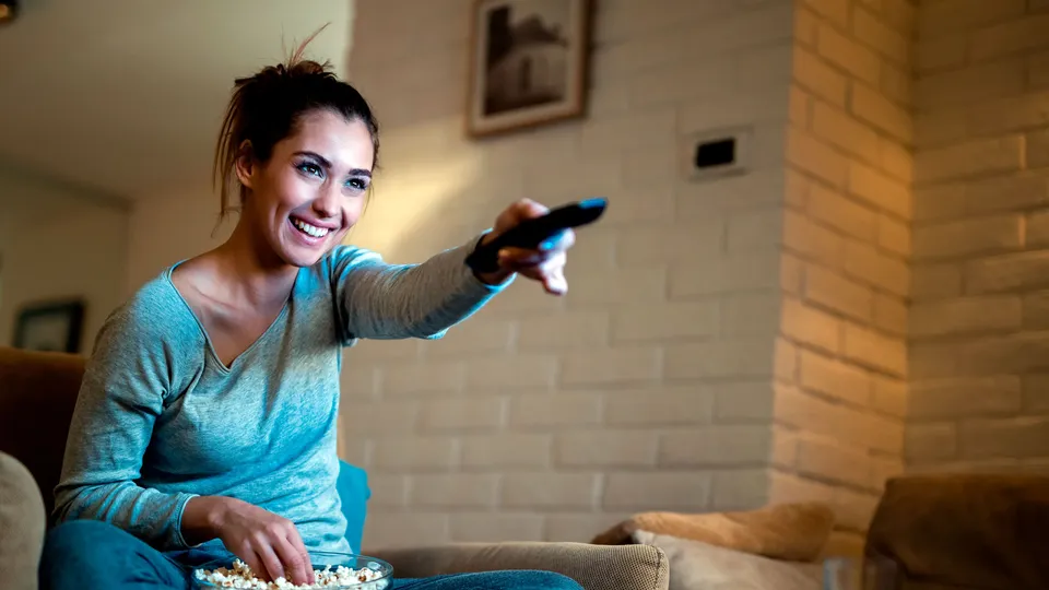 Mujer sosteniendo un control remoto mientras ve televisión con palomitas de maíz.
