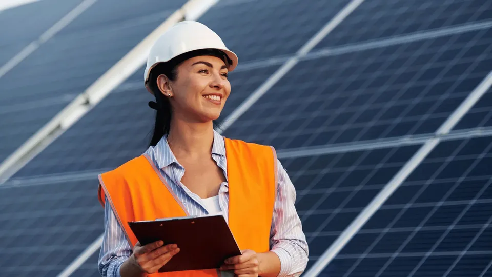 Solar panel maintenance and servicing: Woman in safety helmet and orange vest holding tablet near solar panels.