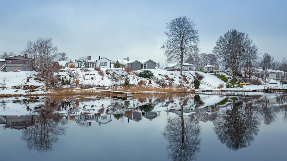Hus och träd vid en spegelblank sjö på vintern. Bryggor går ut i vattnet från hustomterna.